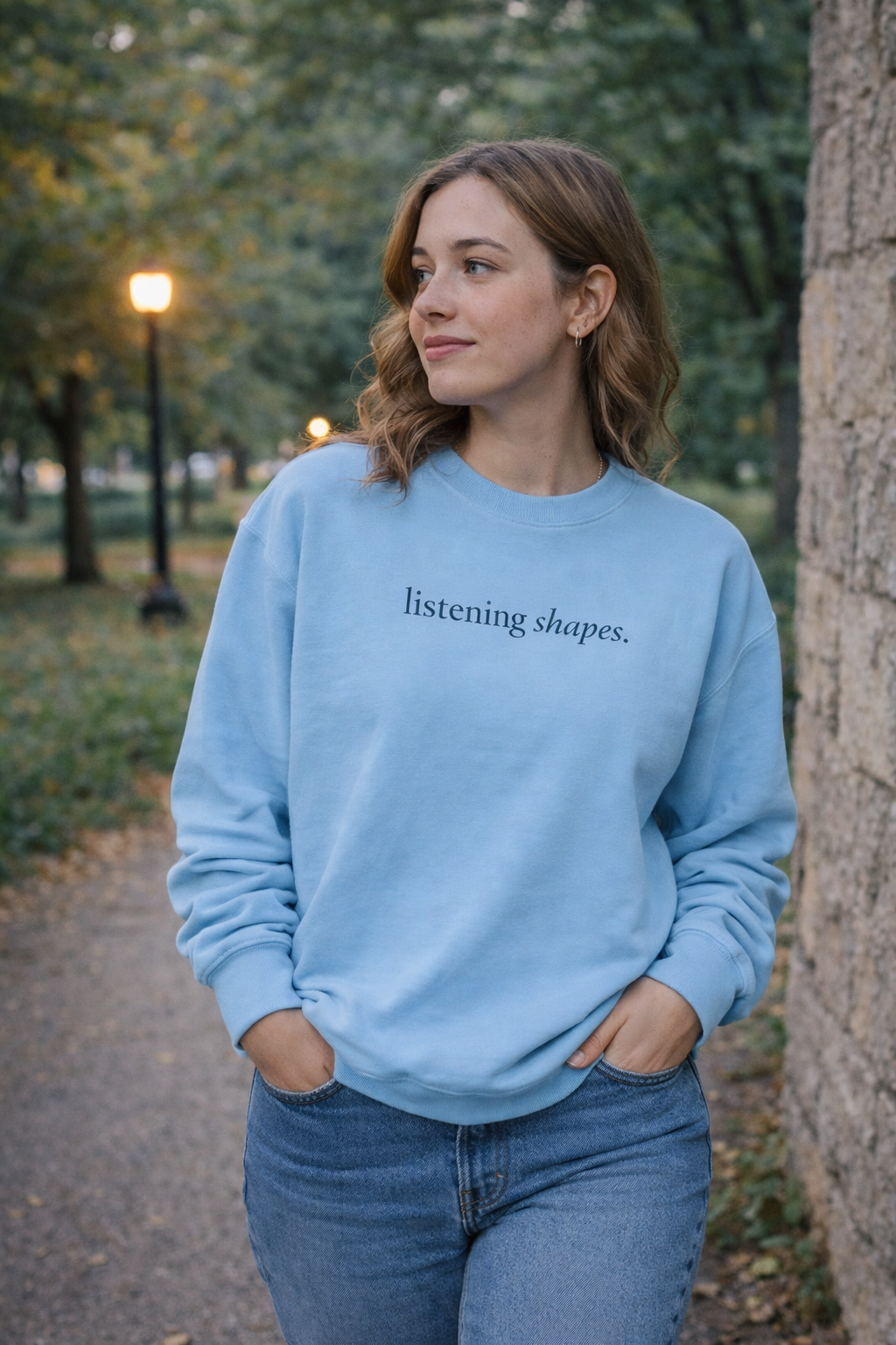 Woman wearing a light blue sweatshirt with 'listening shapes' text, standing outdoors.