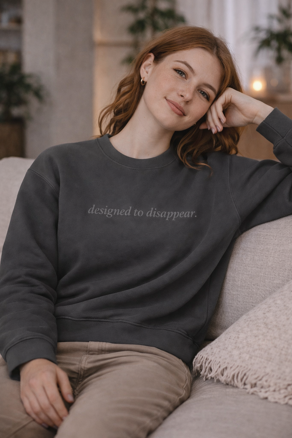 Woman wearing a dark gray sweatshirt with 'designed to disappear' text, sitting on a couch.