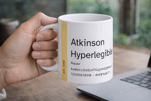 Mug with 'Atkinson Hyperlegible' font design held by a hand on a wooden surface.