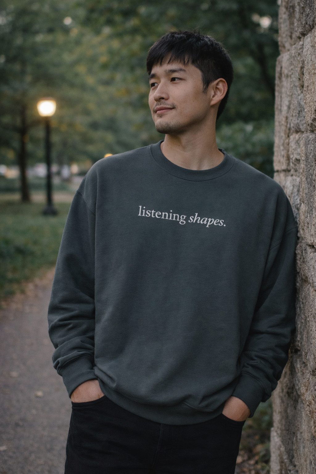 Man wearing a dark gray sweatshirt with 'listening shapes' text, standing against a stone wall outdoors.