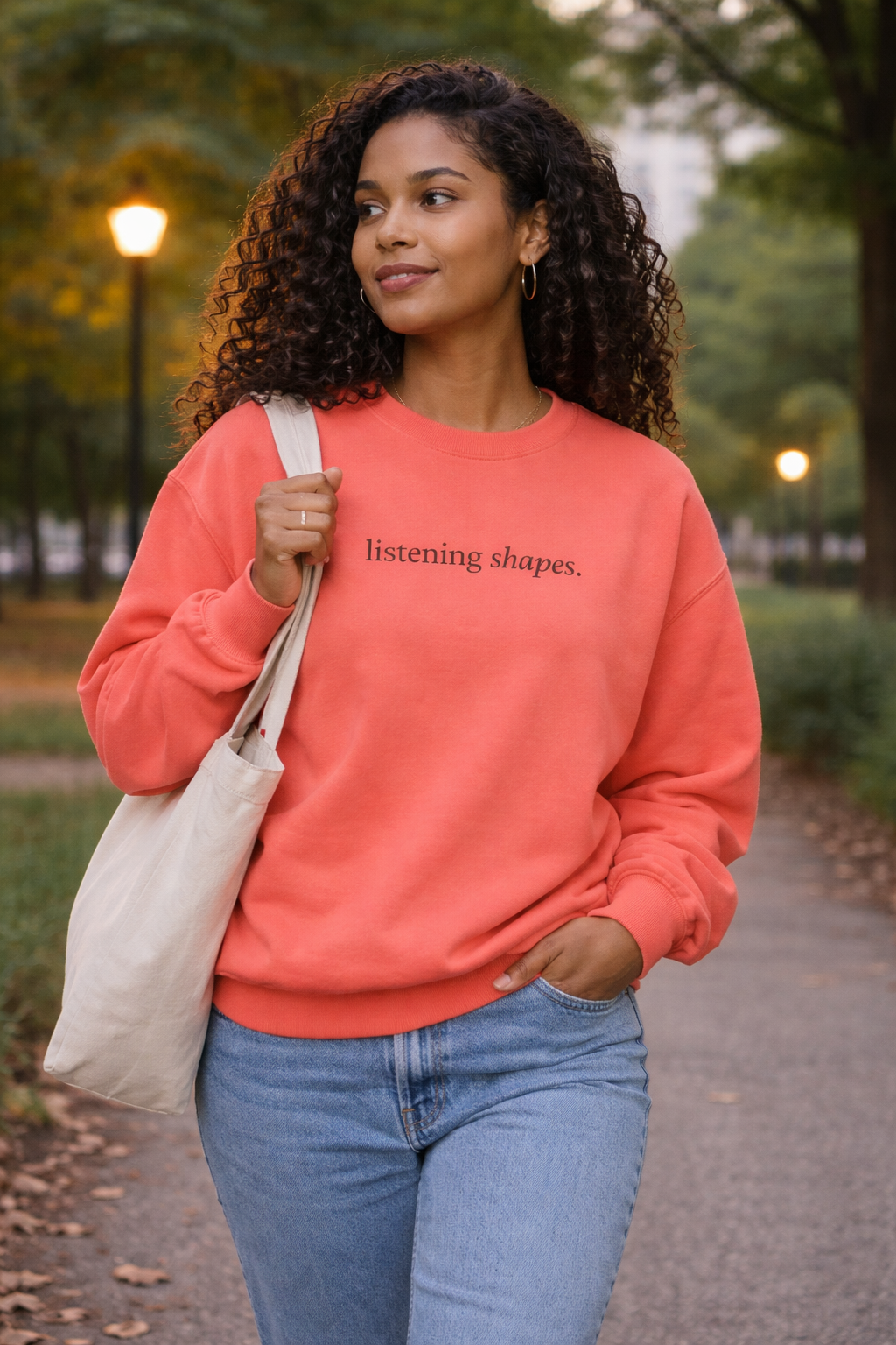 Woman wearing a coral sweatshirt with text, holding a beige bag, standing outdoors.